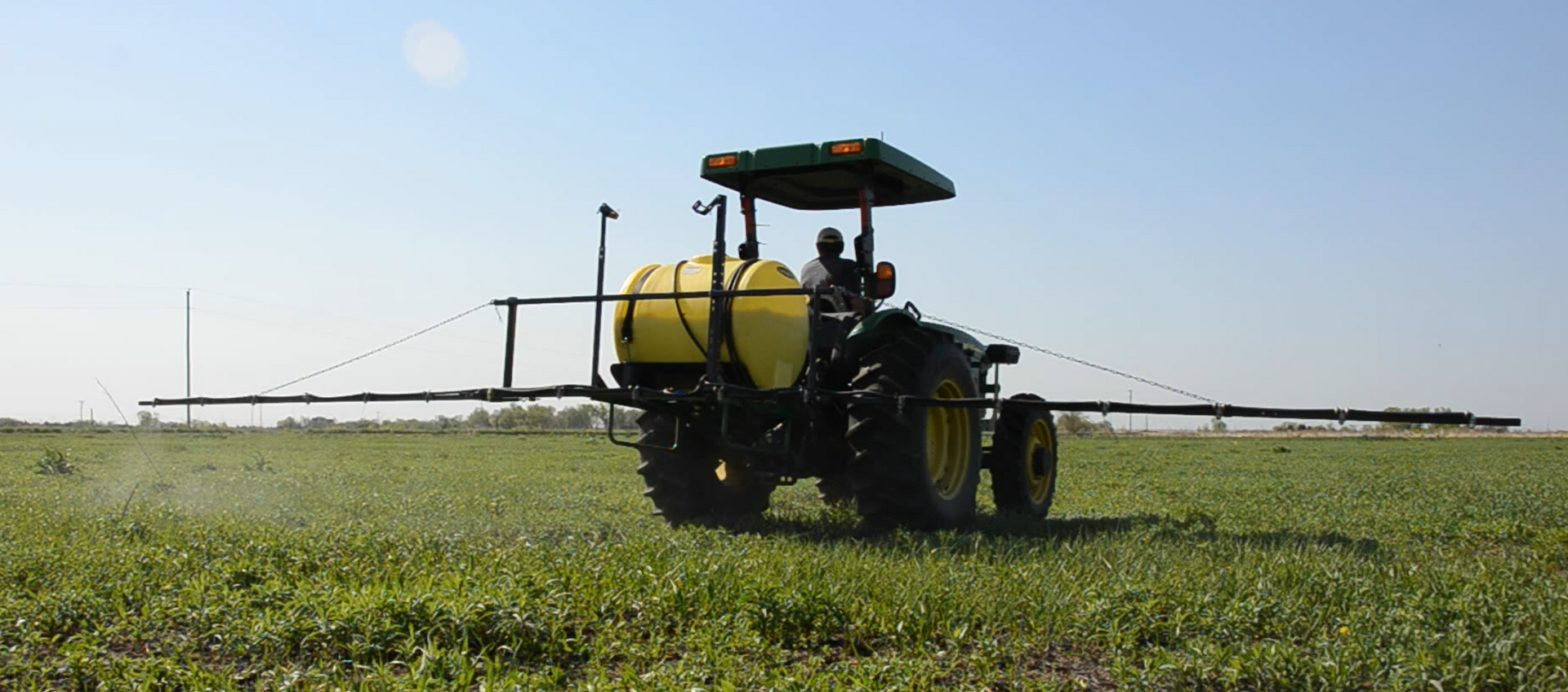 A tractor spraying crops in a green field under a clear blue sky.