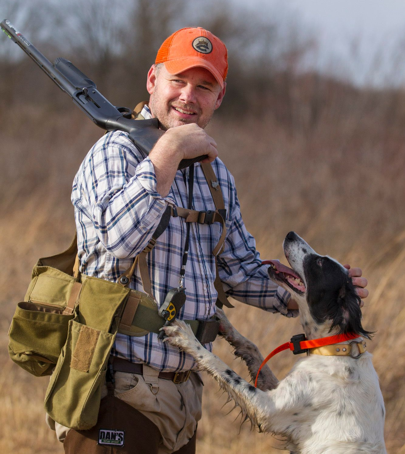 Man in plaid shirt and orange cap with hunting dog in a field. Man holds a shotgun and has a game bag.