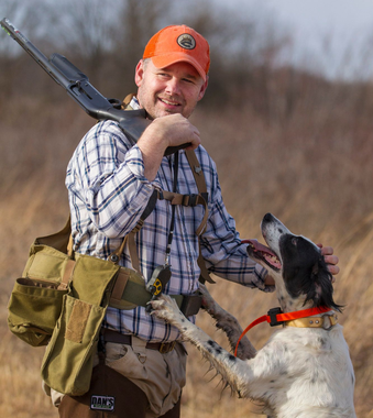 Man in plaid shirt and orange cap with hunting dog in a field. Man holds a shotgun and has a game bag.