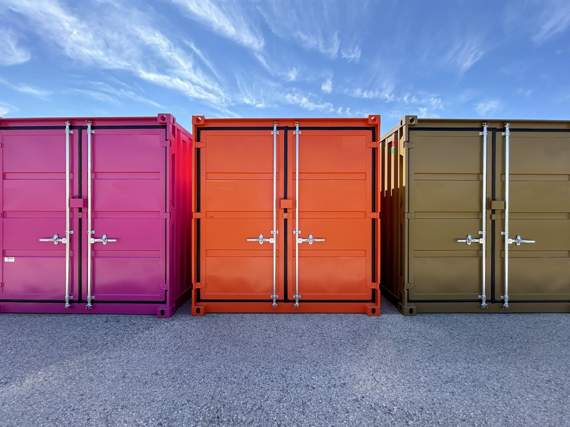 Tre container color viola acceso, arancione brillante e verde oliva sono posizionati uno accanto all'altro su un terreno ghiaioso sotto un cielo azzurro.