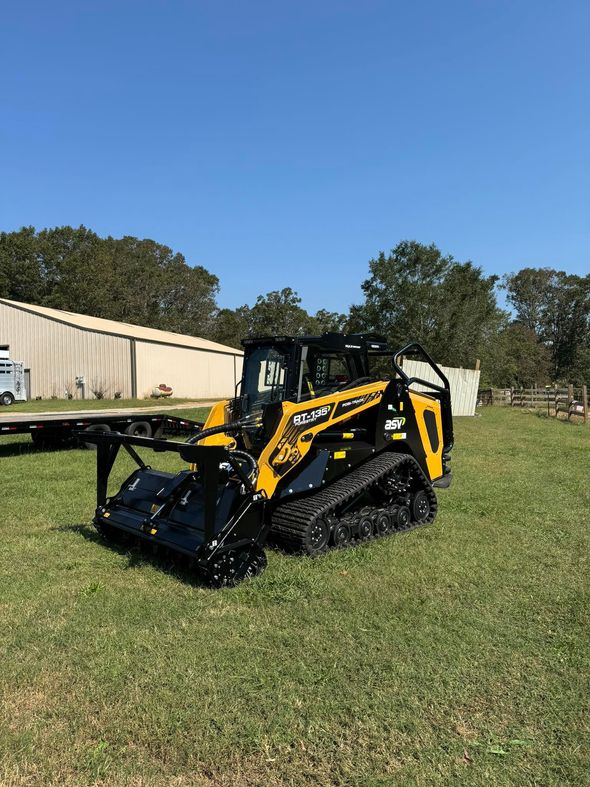 Yellow and black skid steer on tracks with a brush cutter attachment on a grassy field.