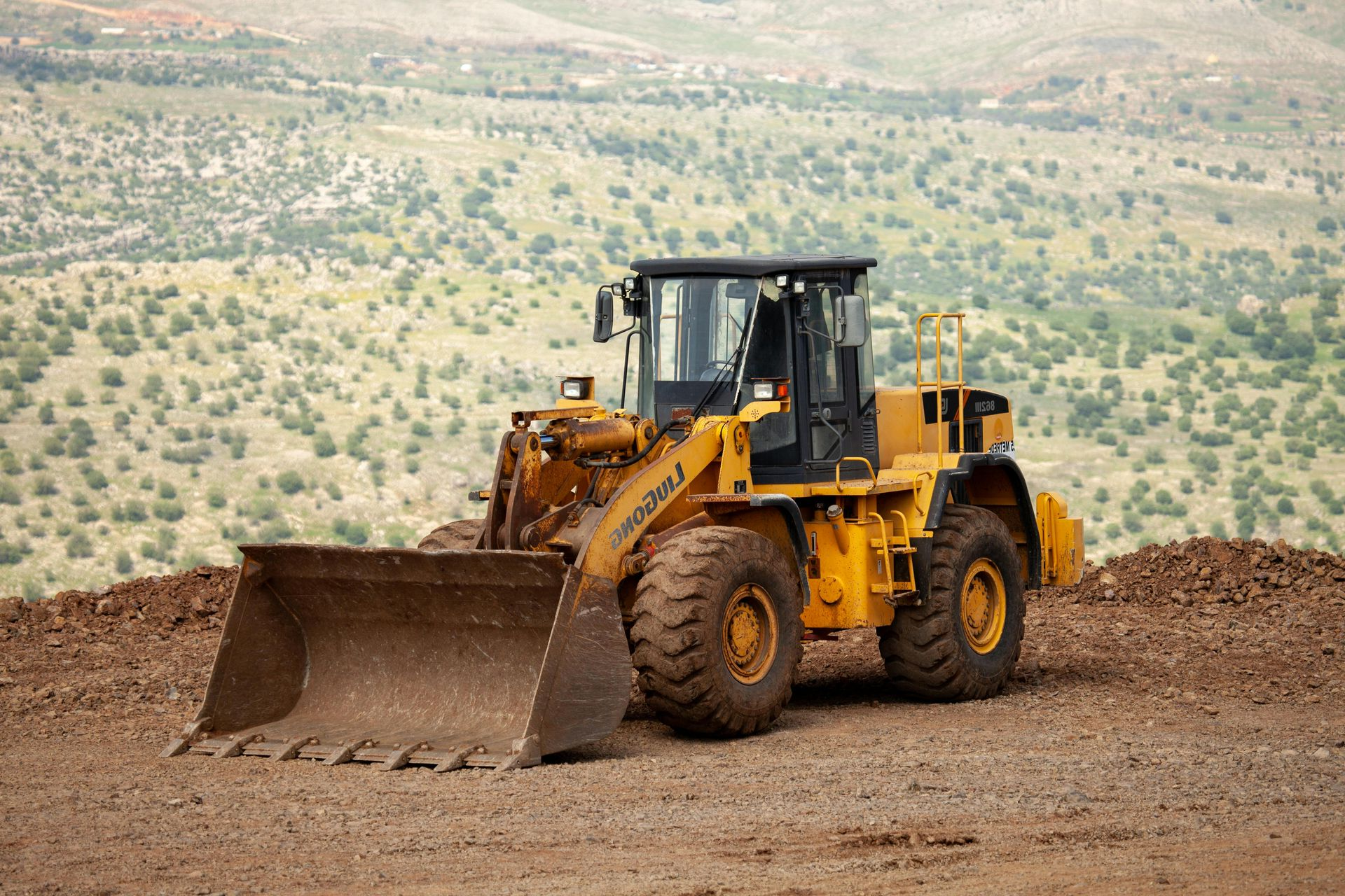 Yellow front-end loader on dirt in front of a hillside with sparse vegetation.
