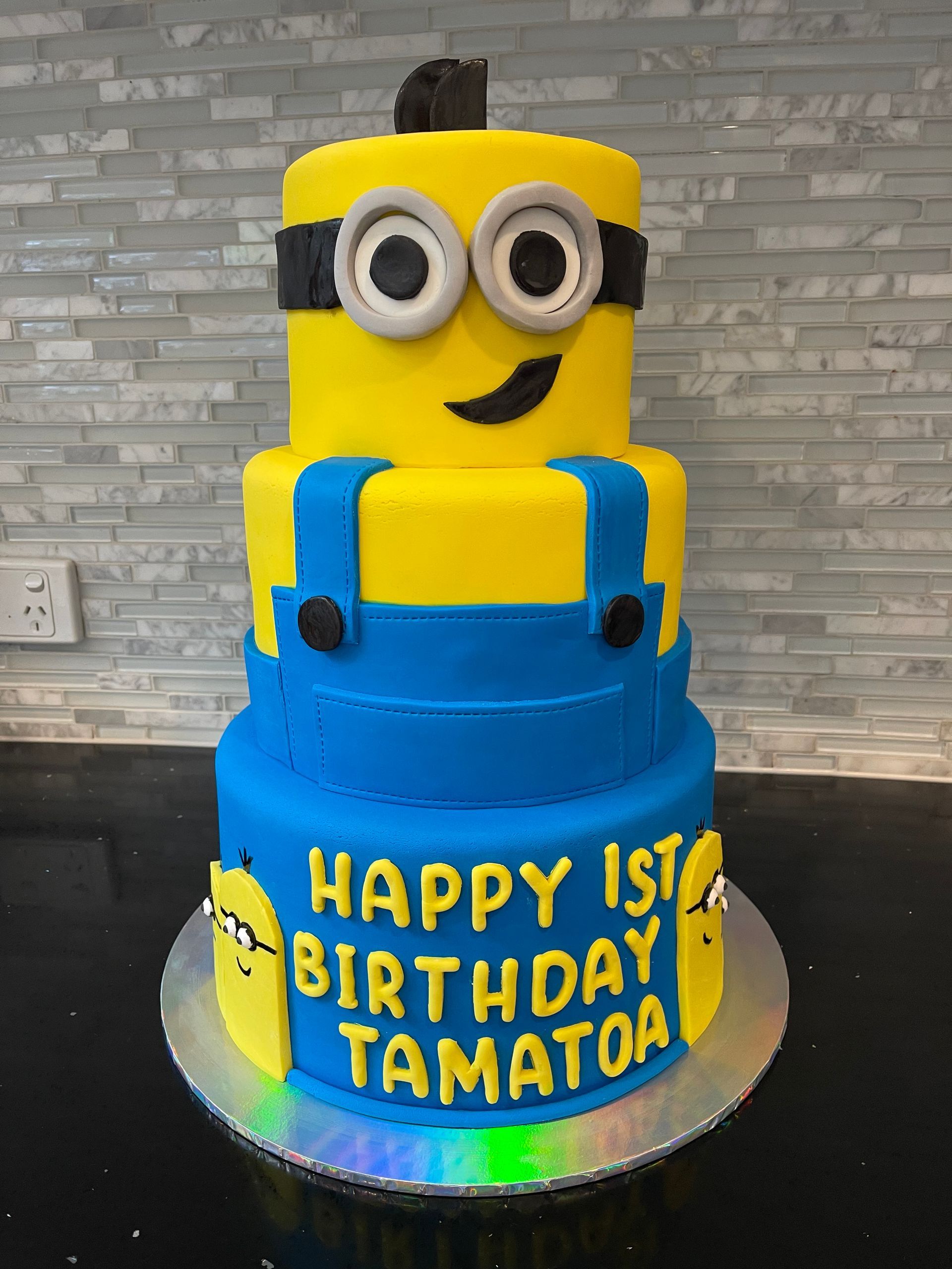 A blue and yellow cake with a minion on top that says happy 1st birthday tamatoa