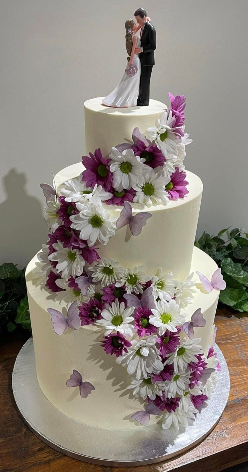 A wedding cake with purple and white flowers and a bride and groom figurine on top.