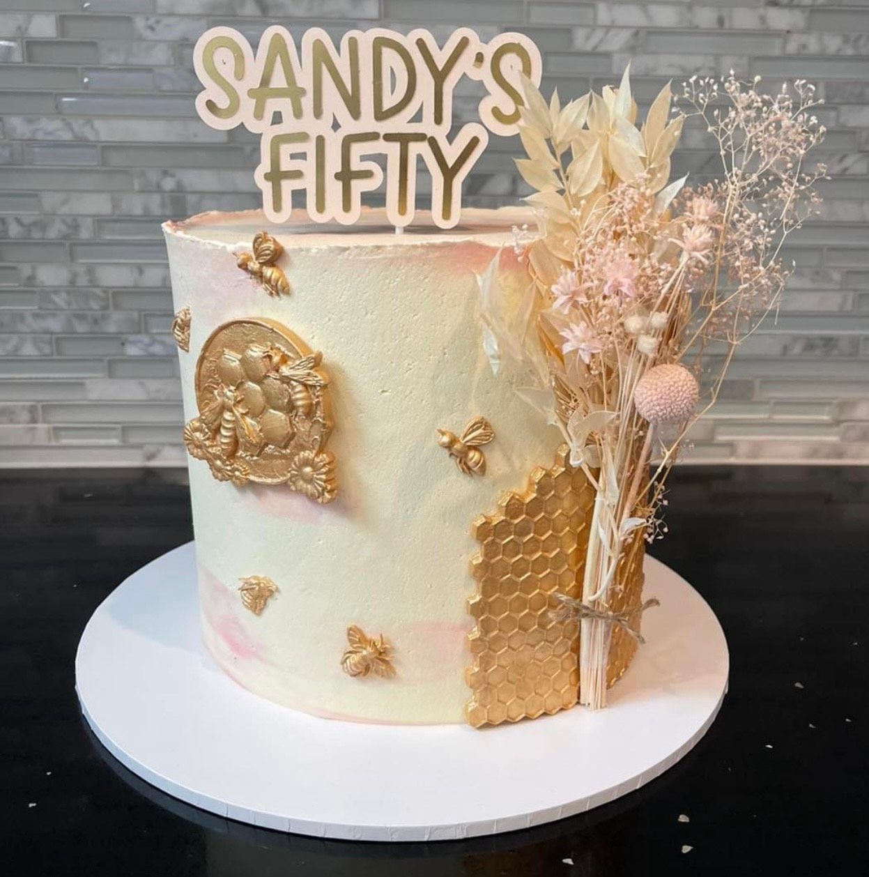 A white cake with gold decorations and a sign that says sandy 's fifty