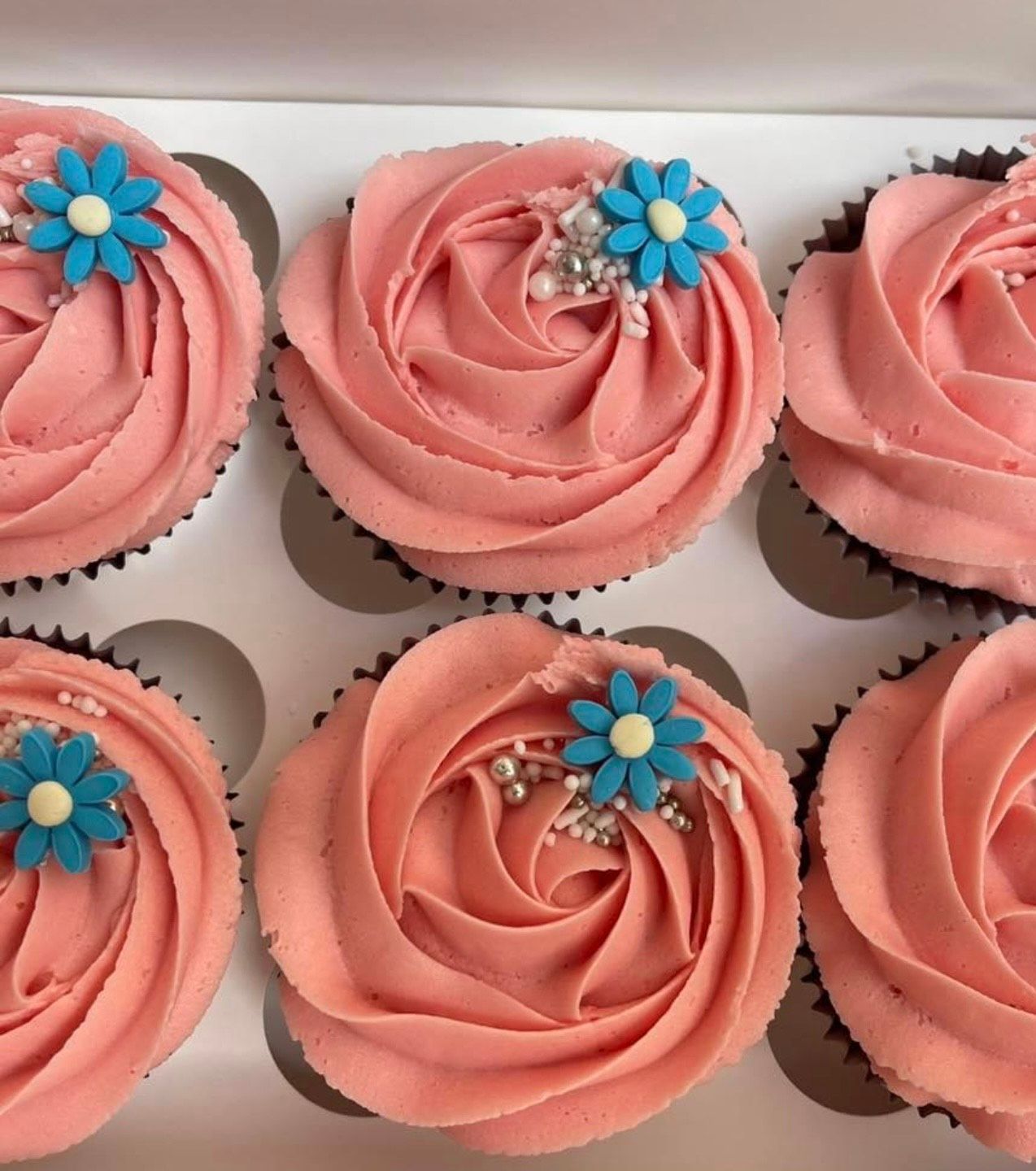 A box of pink frosted cupcakes with blue flowers on top