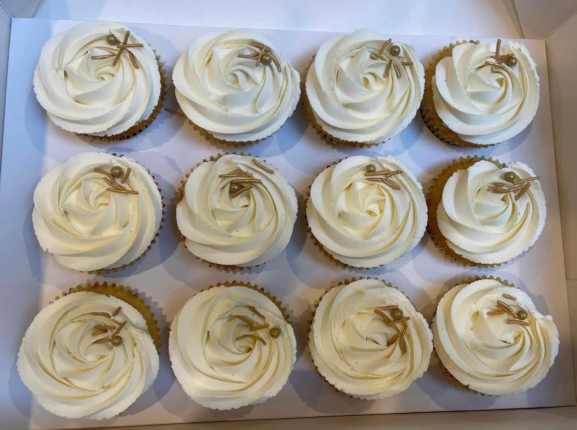 A box of cupcakes with white frosting and gold sprinkles.