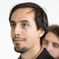 A man with a beard is looking at the camera with a woman in the background.