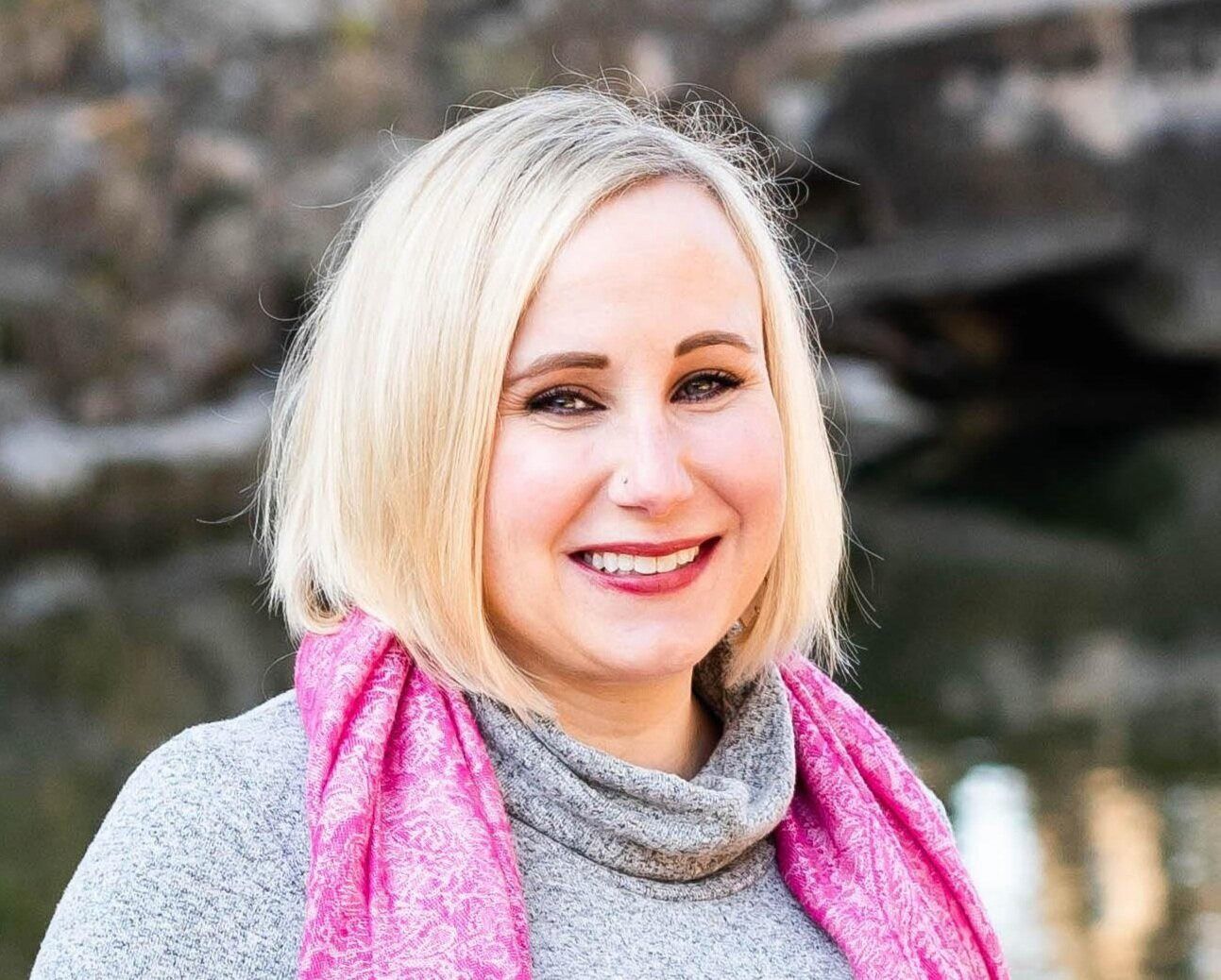 A woman wearing a gray sweater and a pink scarf is smiling for the camera.
