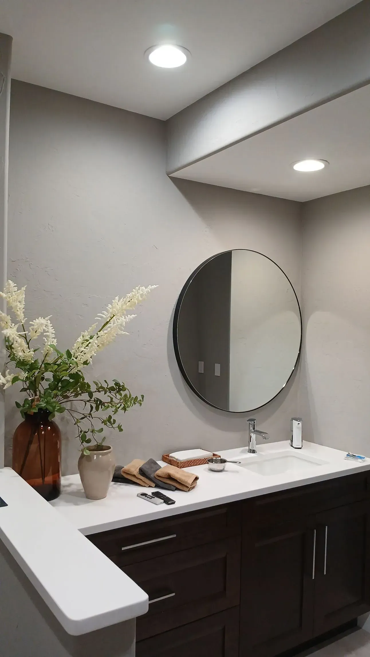 Bathroom with dark wood vanity, round mirror, white countertop, and vase of flowers.
