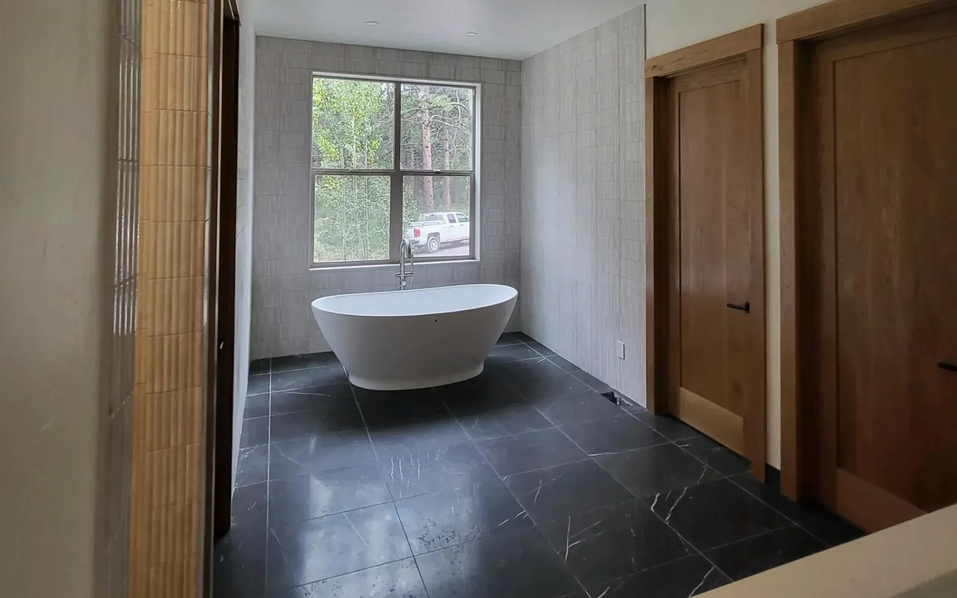 Modern bathroom with white tub, large window, dark floor, and wood doors.