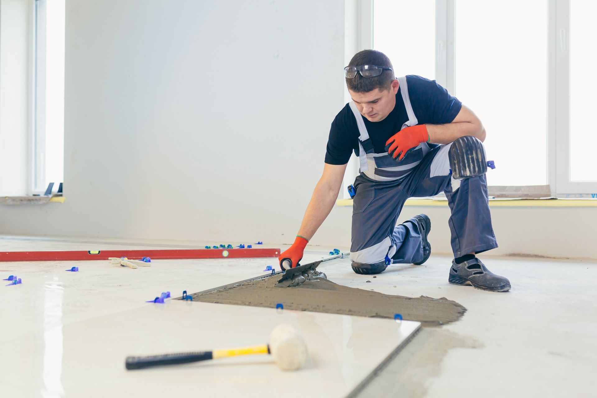 Man kneeling on floor applying mortar to tile. Construction site interior, tools visible.