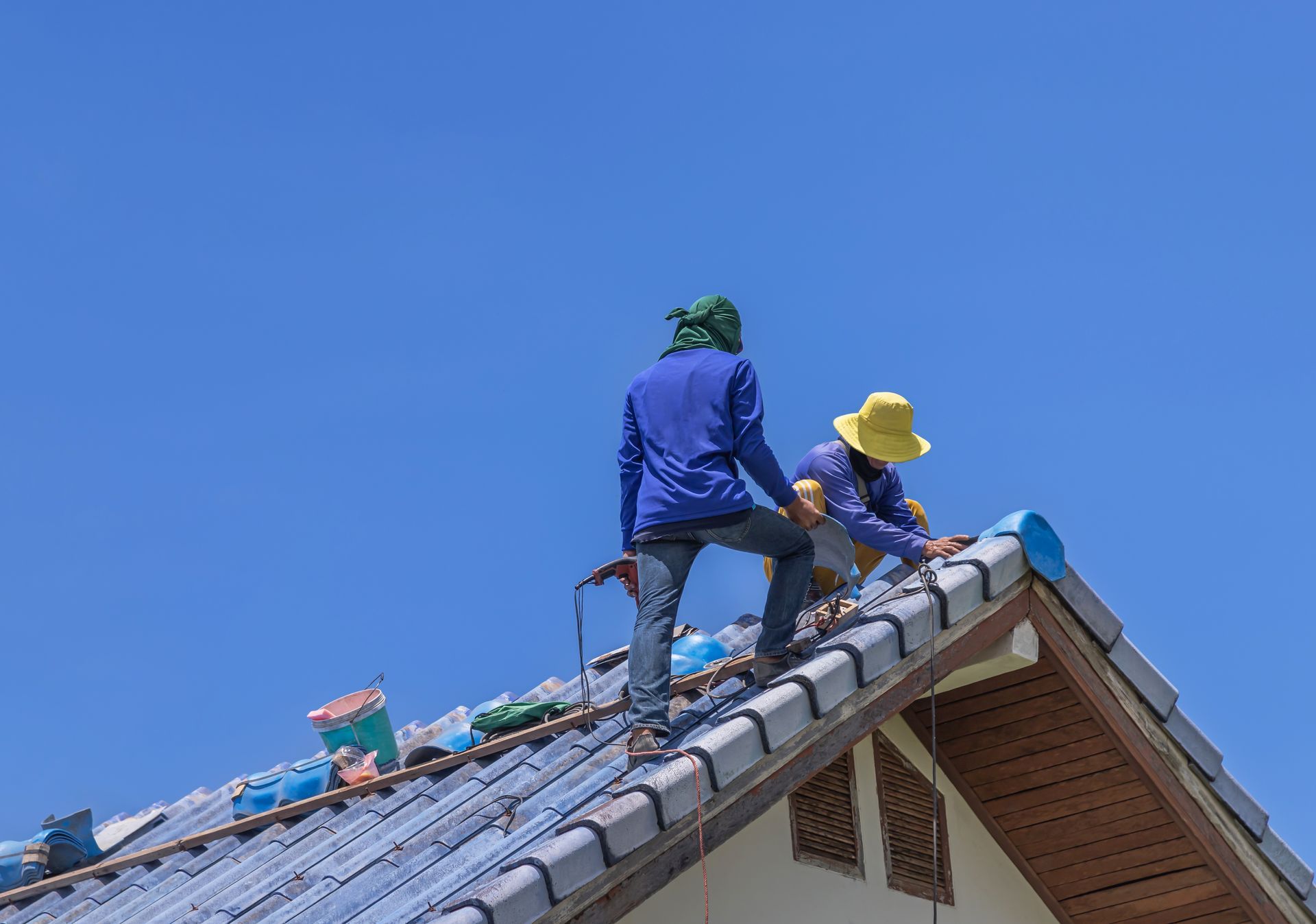 Two workers installing blue roof tiles on a house under a clear, blue sky.
