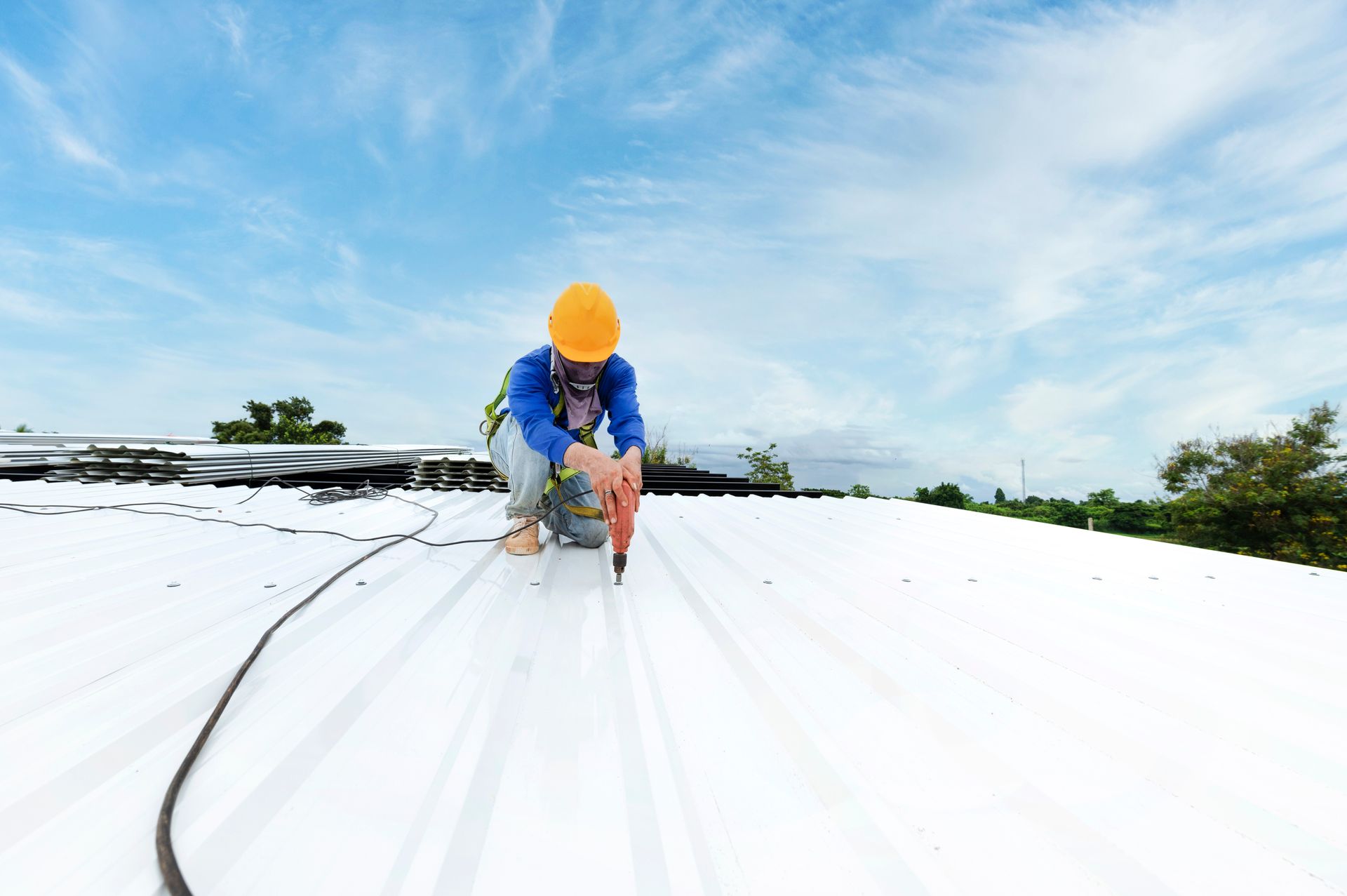 Roofer in yellow hard hat, using a tool on a white metal roof under a blue sky.