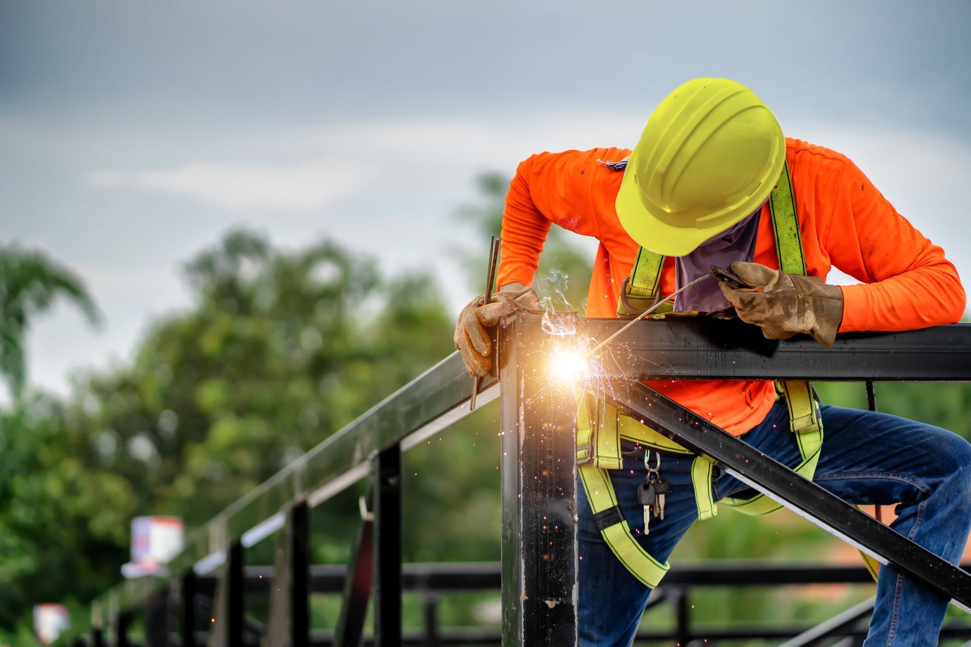 Welder in orange shirt and yellow hard hat welding a metal railing outdoors.