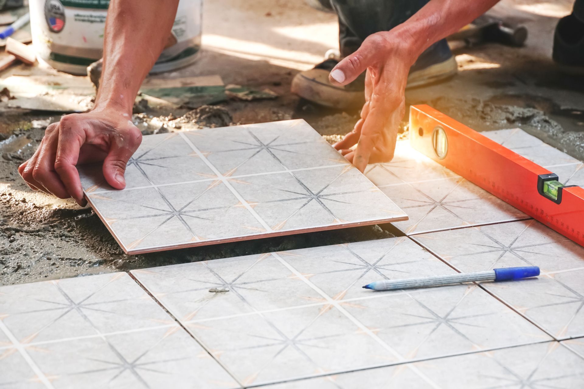 Hands laying a patterned tile on a concrete surface, with a level and marker nearby.