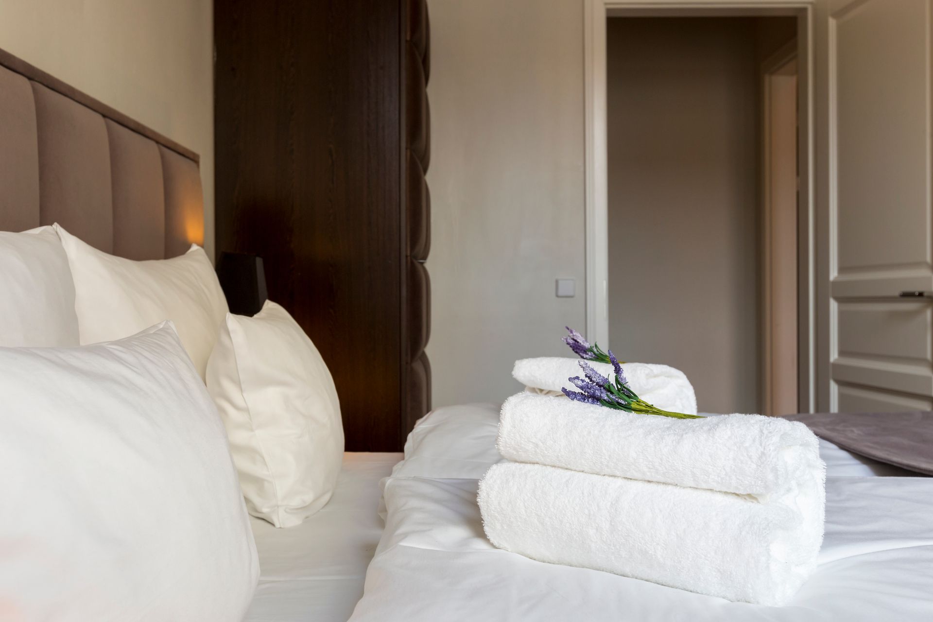 Bedroom with white bedding, stack of towels, sprig of purple flowers, closet, and open doorway.