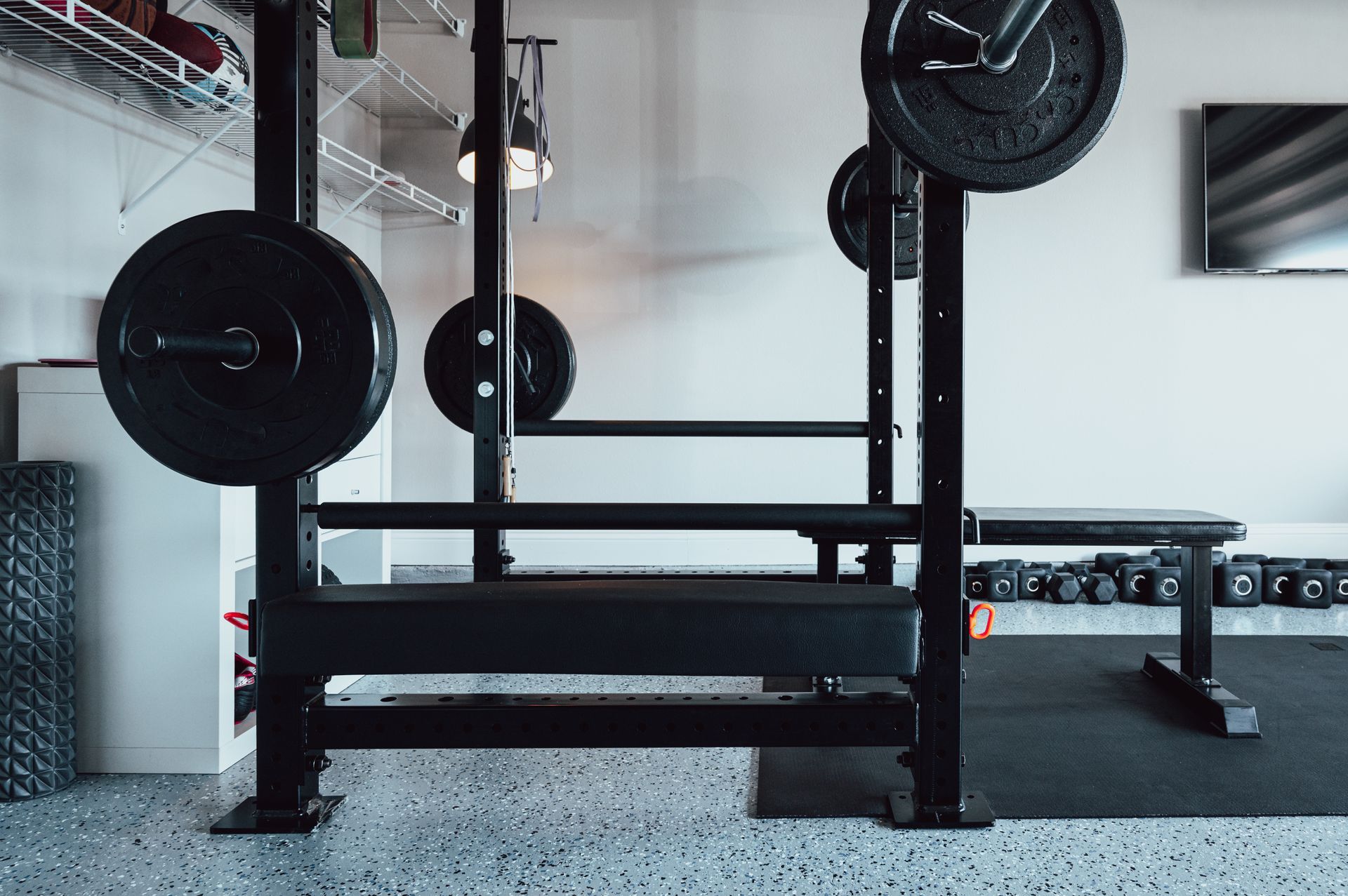 Weightlifting equipment in a home gym. A black barbell rack with weights, bench, and free weights on the floor.