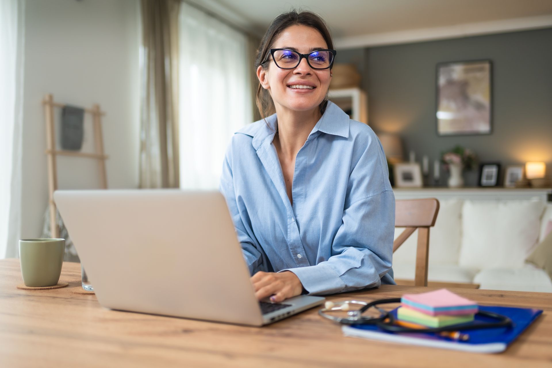 Woman in glasses smiles while working on laptop at a wooden table in a well-lit room.
