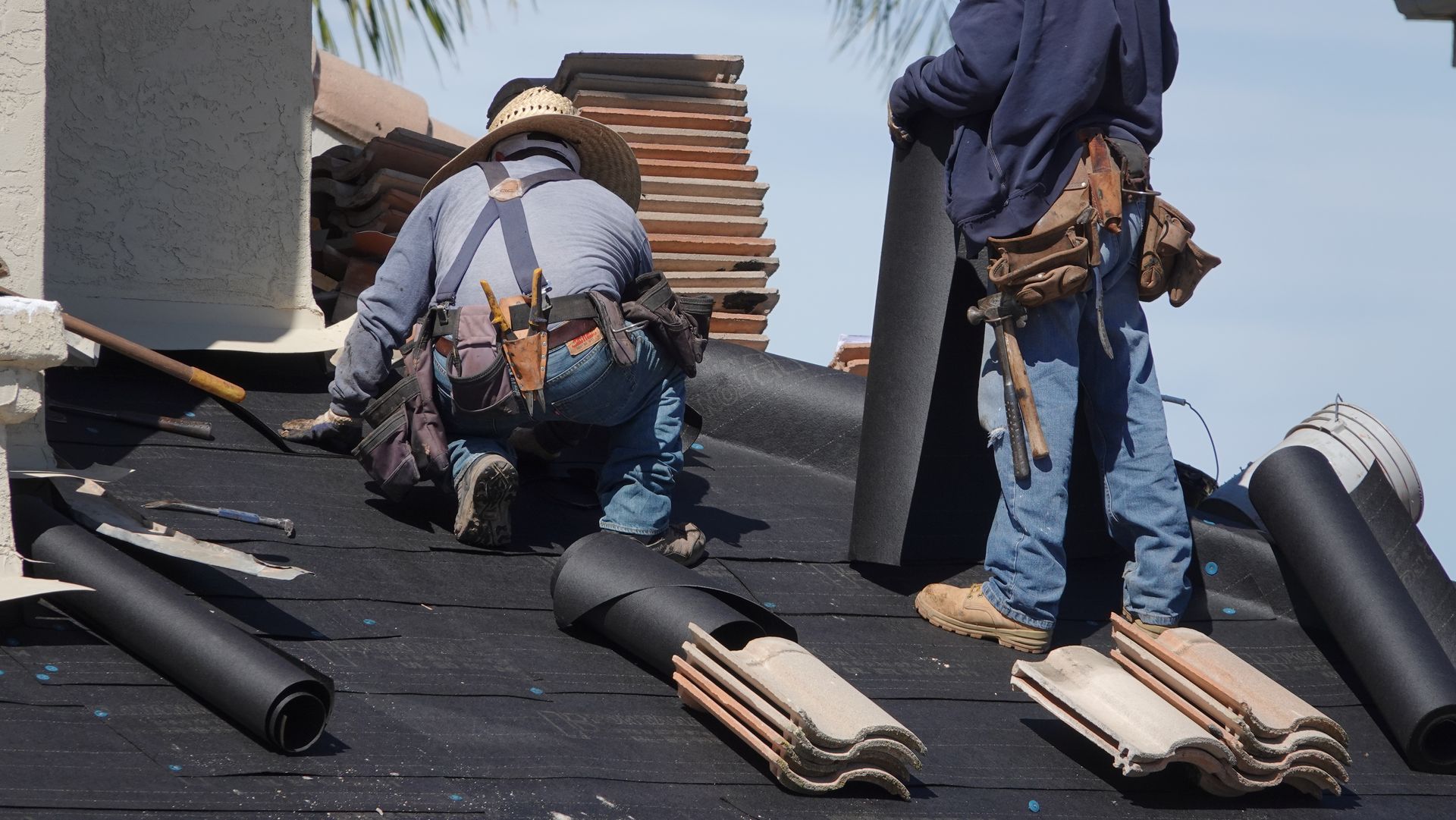 Person in work clothes using a drill to install roof boards on a wooden structure outdoors.