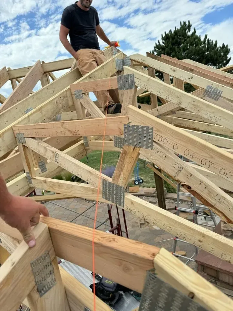 Man working on a wooden roof frame under a partly cloudy sky.