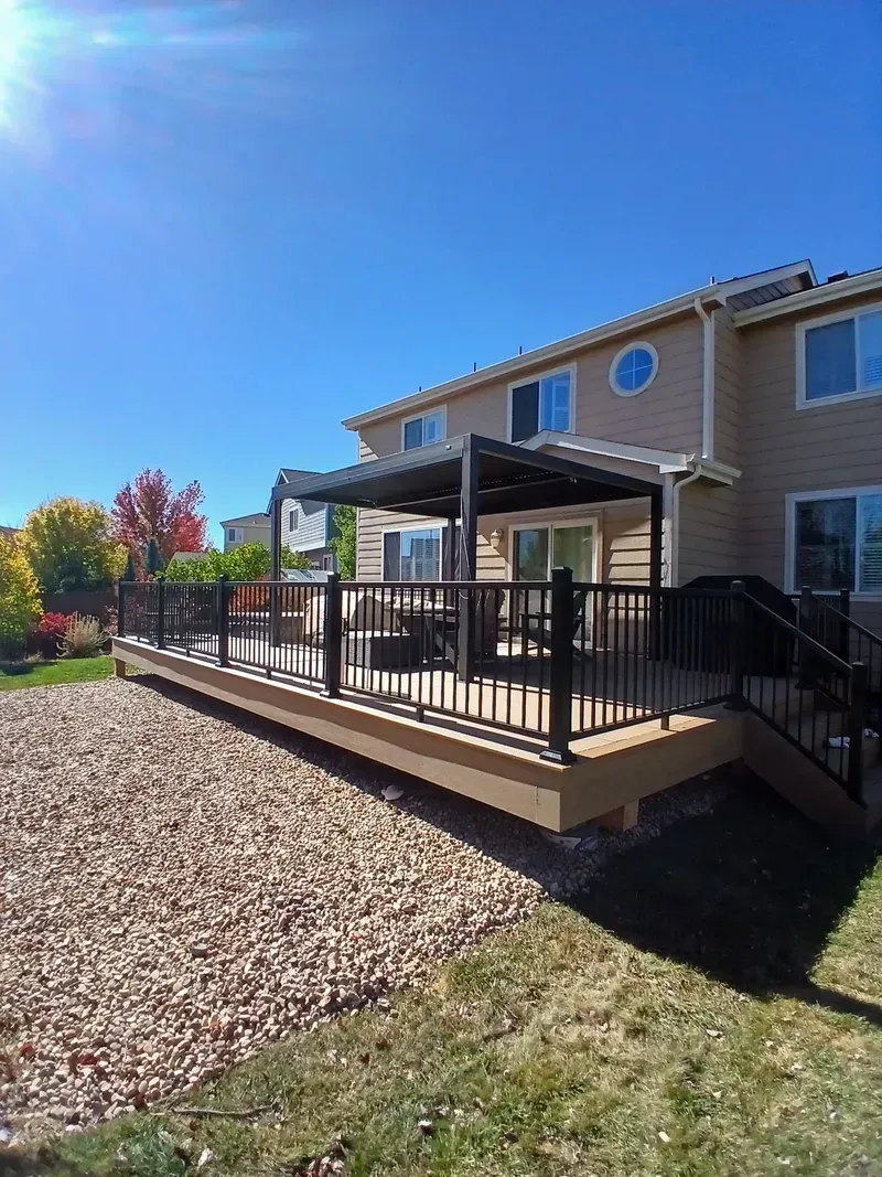 Backyard deck with pergola, overlooking a rock-covered slope and green grass, house in background, blue sky.