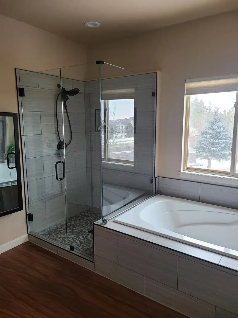 Bathroom with glass shower, white tub, and tile. Brown flooring, and a window with outside view.
