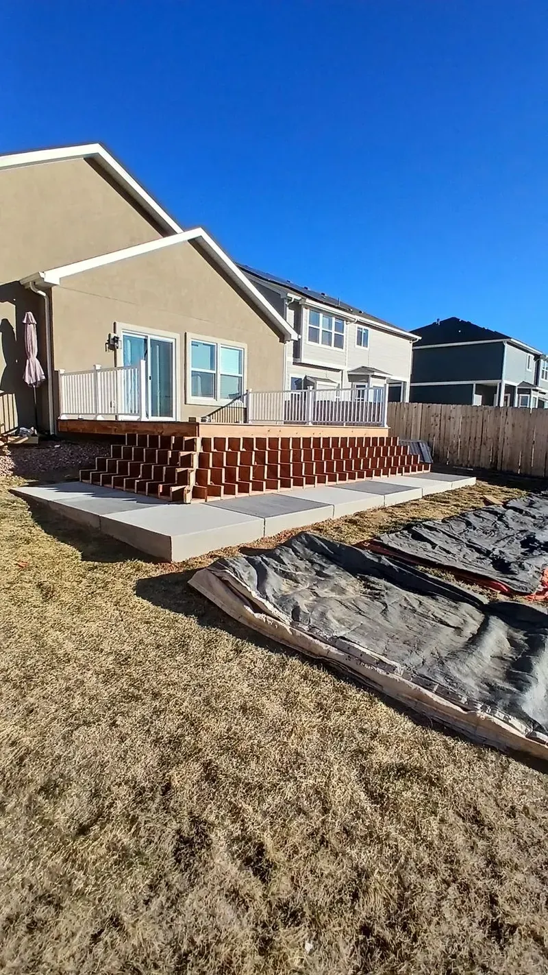 Backyard deck under construction; brown wooden frame, attached to a light-colored house, concrete patio, clear blue sky.