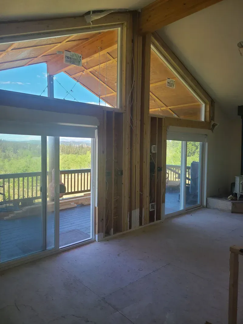 Interior view of a house under construction with large windows and a deck. Wooden beams are visible.