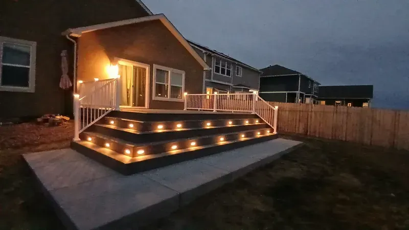 Nighttime view of a multi-level deck with built-in lights and white railings, attached to a house with a concrete patio.