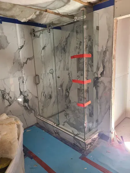 A shower enclosure under construction, featuring marble-look tiles, glass doors, and orange shelves.