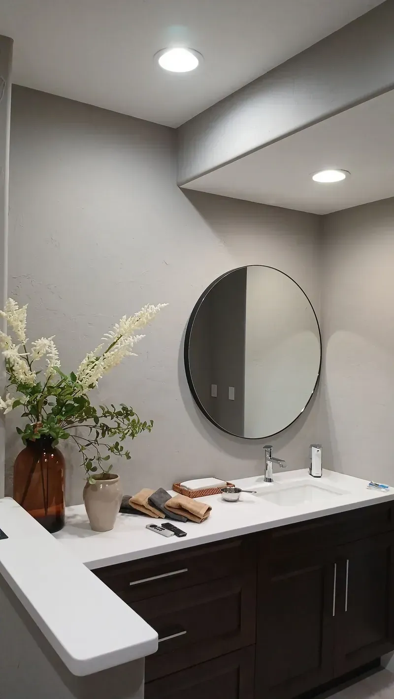 Bathroom with dark brown vanity, round mirror, white countertop, and flowers.