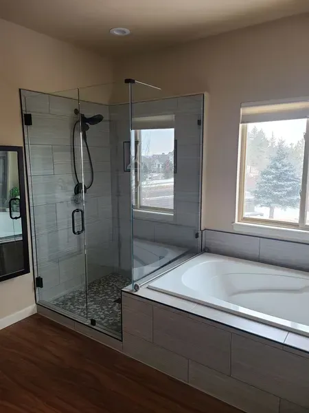 Bathroom with glass shower, white tub, window, and wood-look flooring.