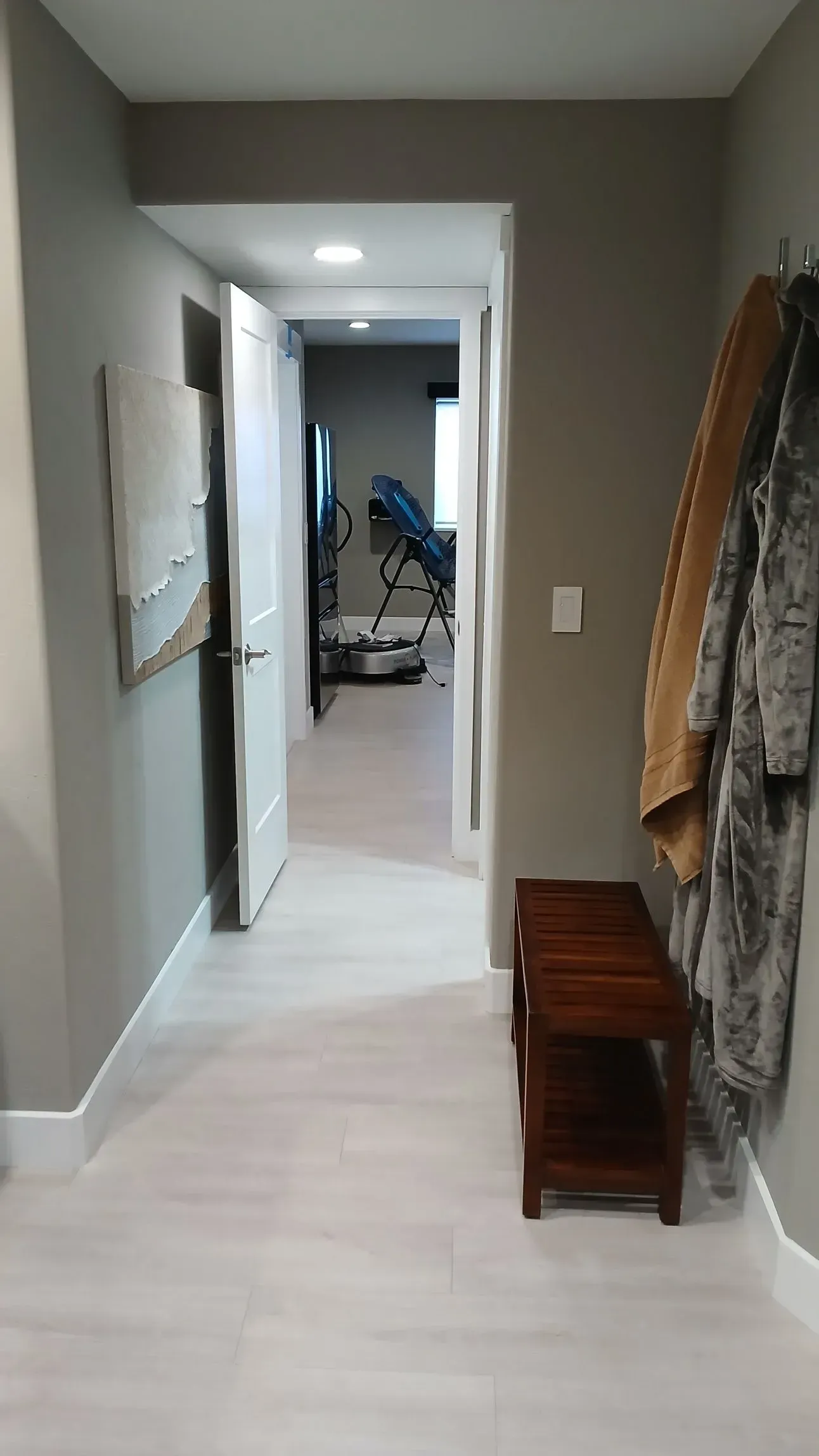 Hallway with light wood floors, gray walls, open door, bench, and coats hanging.