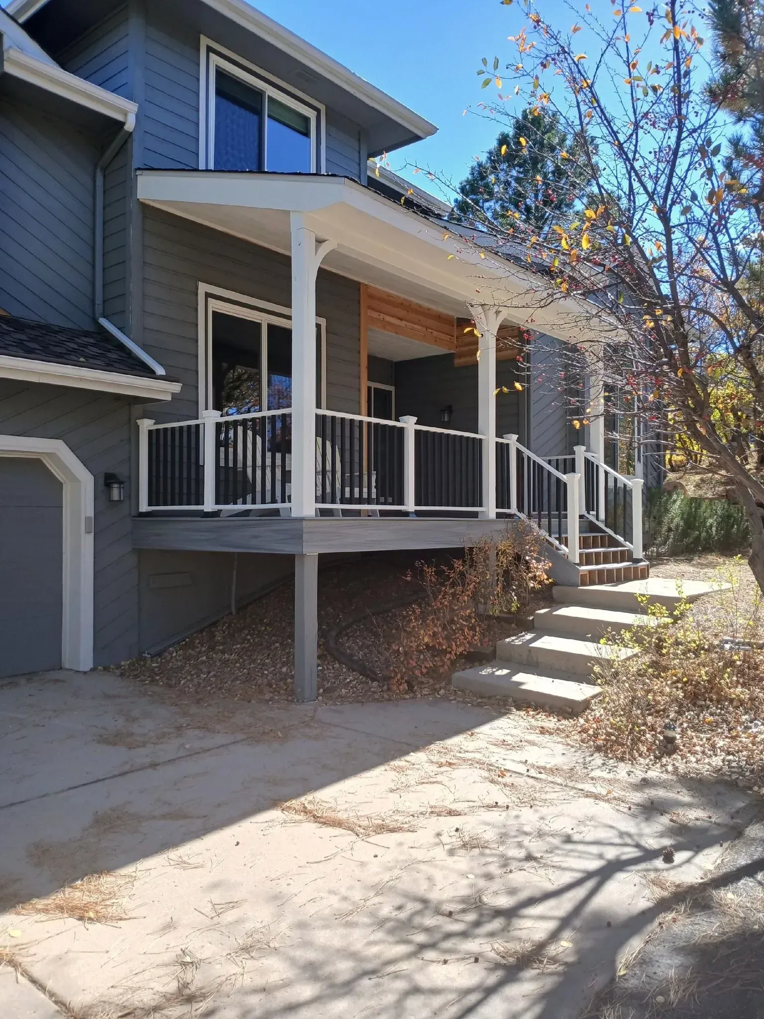Two-story gray house with a porch and deck. Stairs lead up to the deck. Light falls on the concrete driveway.