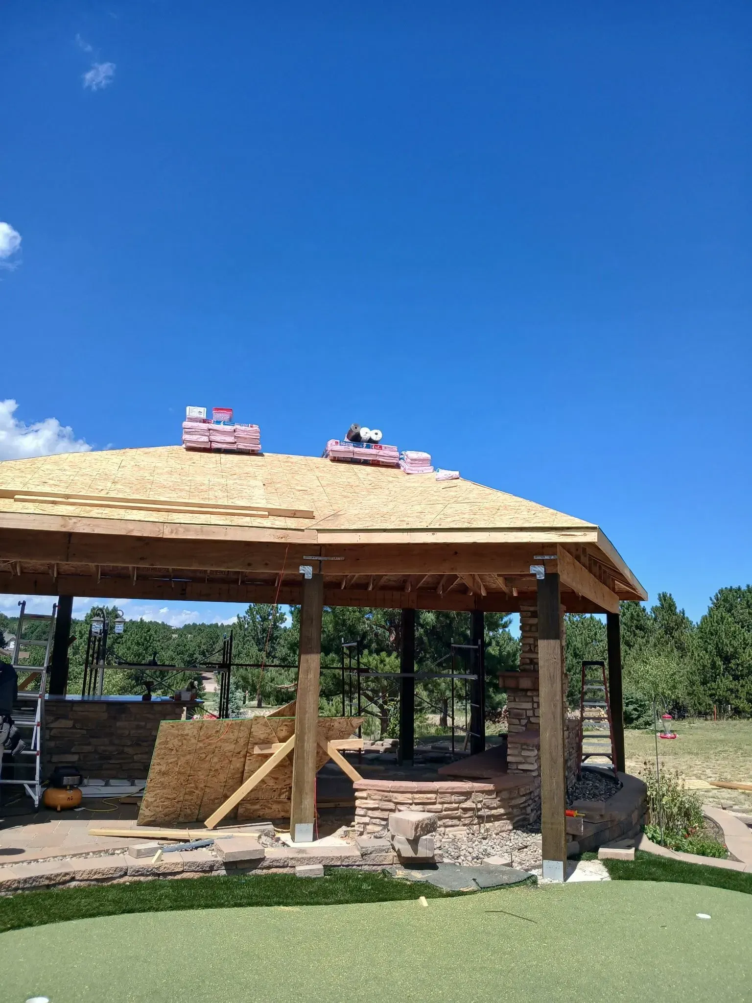 A partially constructed outdoor pavilion under a blue sky, with roofing materials on top.