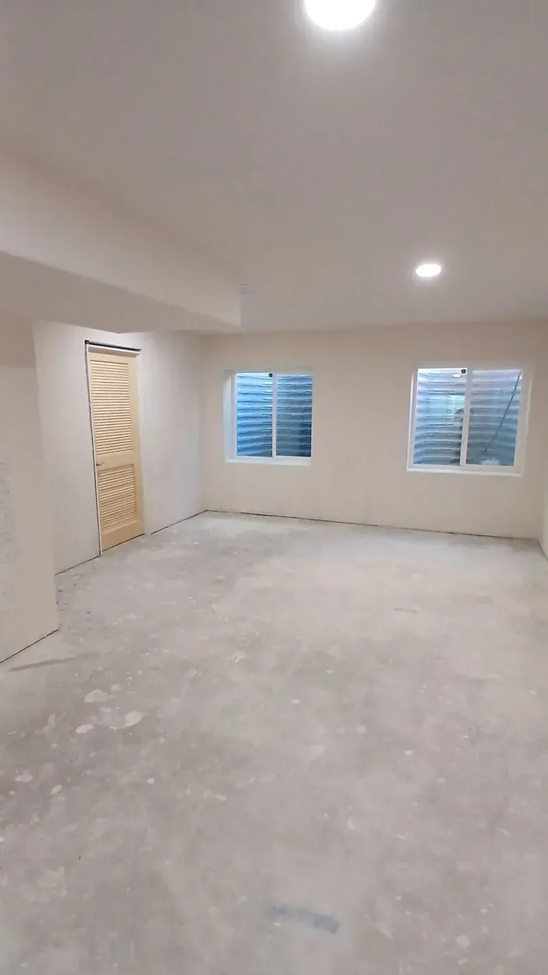 Empty unfinished basement room with two windows and a door. Light gray walls and floor.