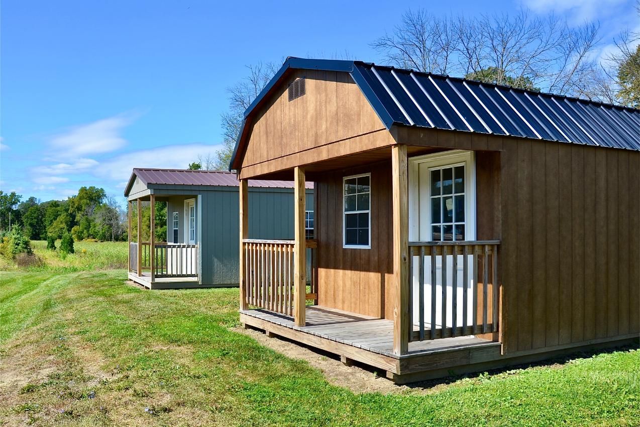 Two wooden shed-style tiny houses with front porches sit in a grassy field under a bright blue sky.