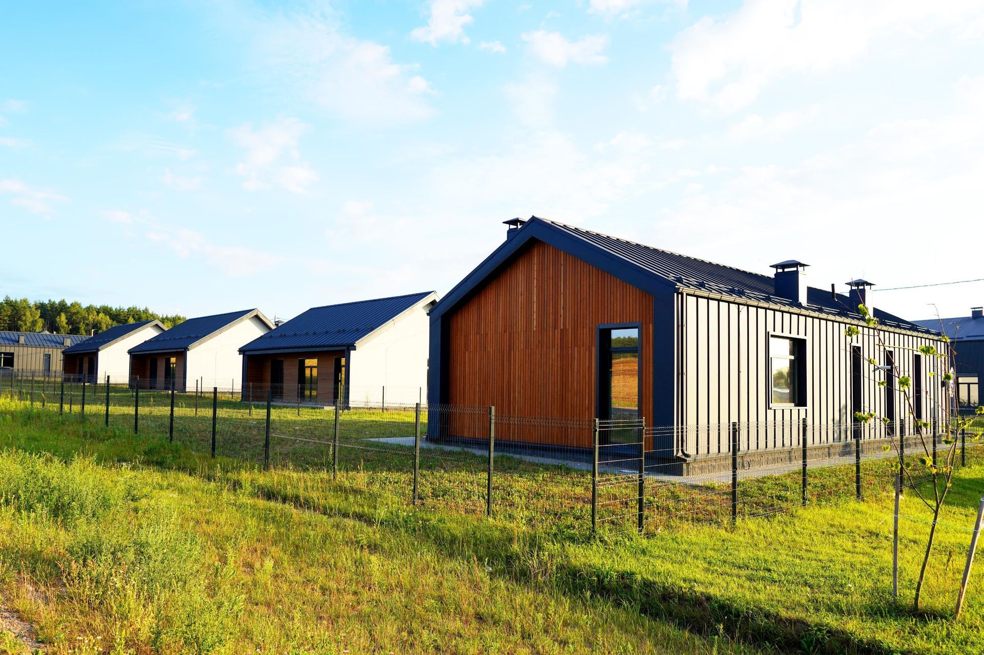 A row of modern suburban houses with wooden and dark metal siding, seen from a grassy field under a bright blue sky.