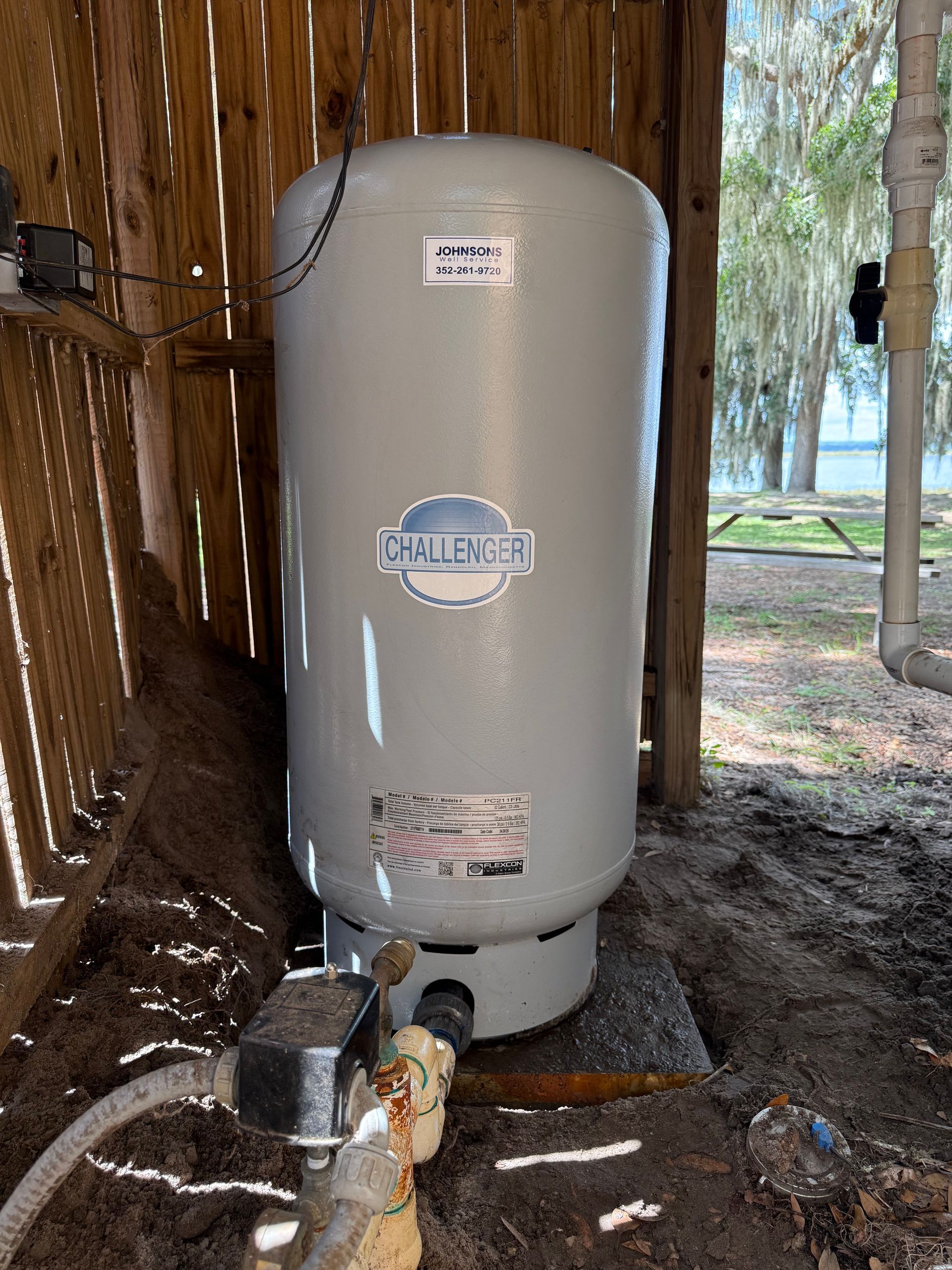 Water pressure tank inside a wooden shed, next to pipes and a grassy outdoor view.