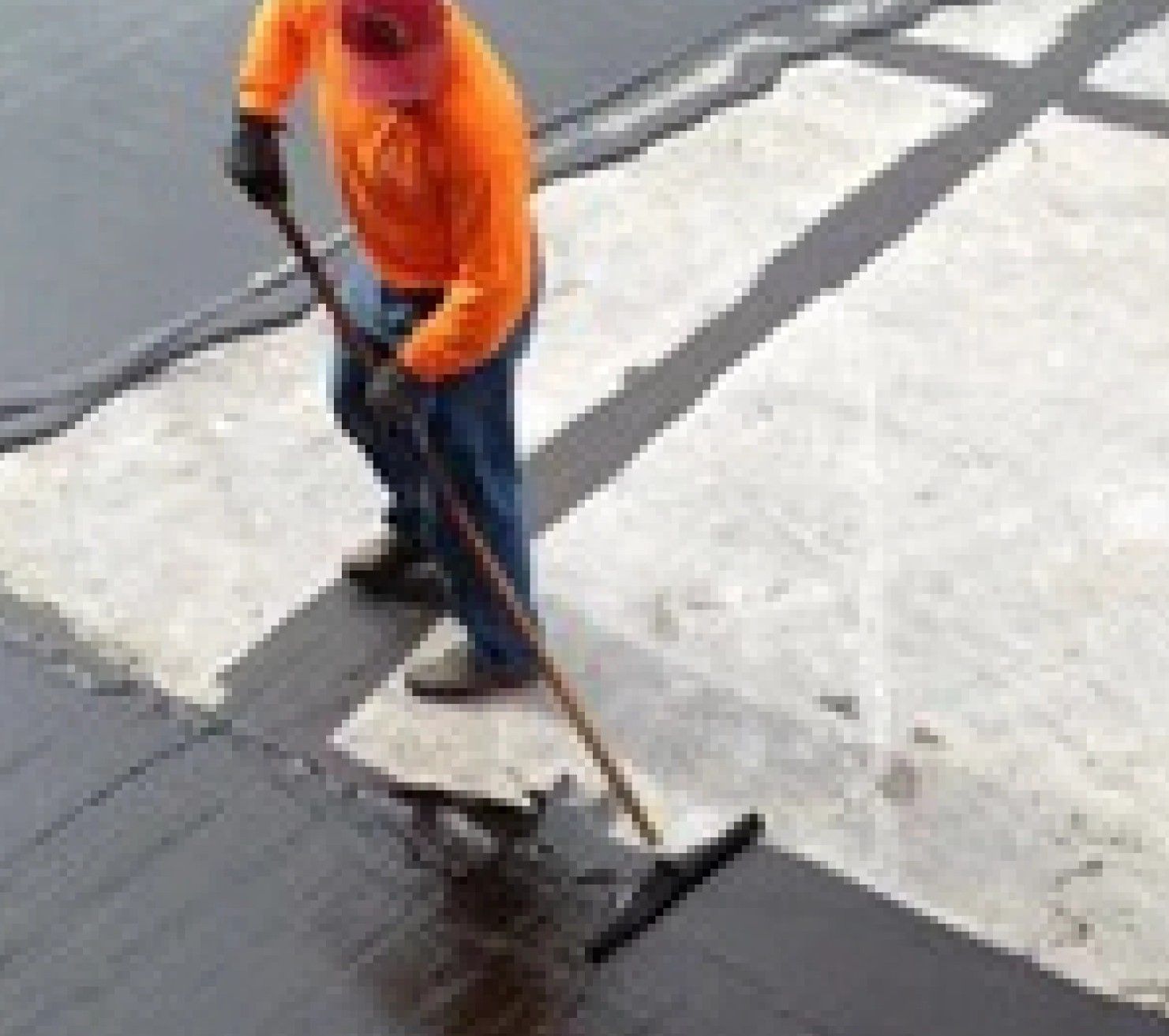 A man in an orange shirt is using a broom to clean a street.