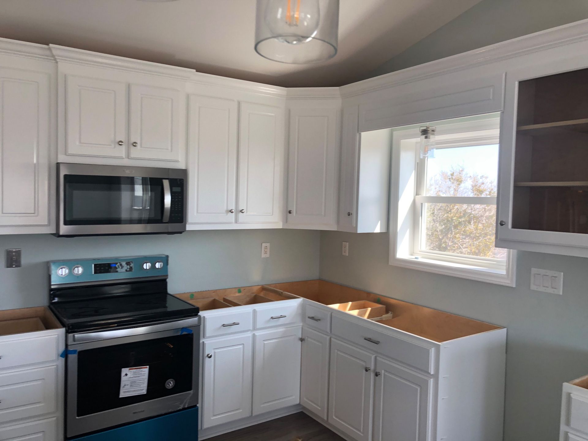 A kitchen with white cabinets , a stove , a microwave and a window.