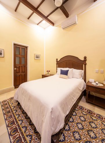 Dormitorio con cama de madera, mesitas de noche y alfombra estampada. Paredes amarillas y techo con vigas de madera.