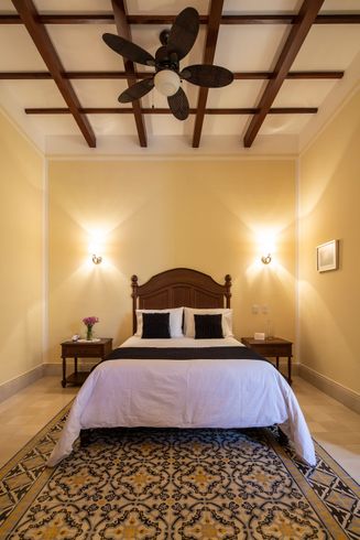 Dormitorio con cabecera de madera, mesas de noche y ventilador de techo; piso de baldosas estampadas, paredes color crema.