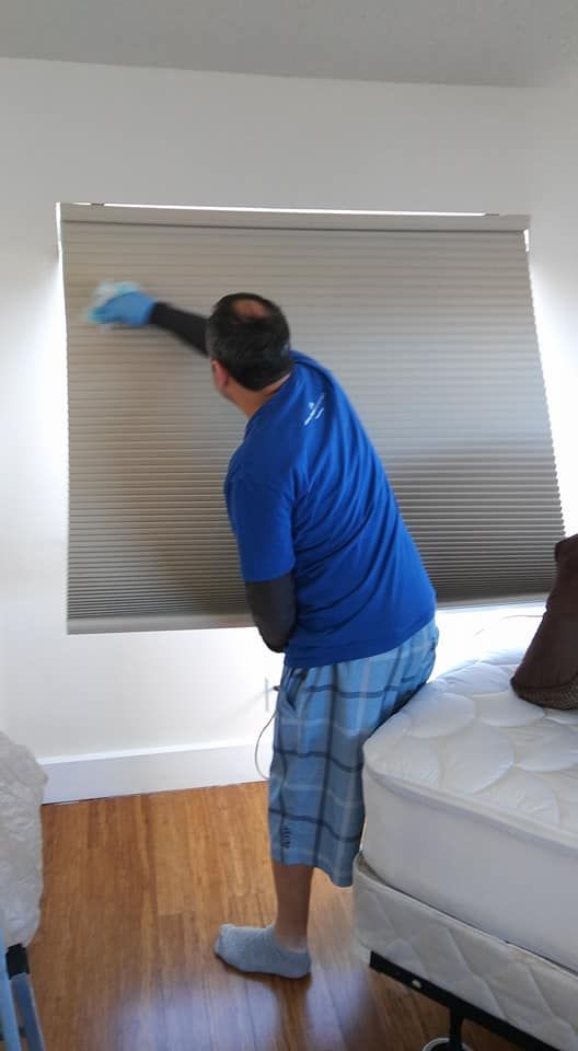 A man is cleaning a window blind in a bedroom.