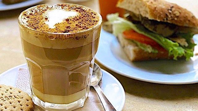 A cup of coffee and a sandwich on a plate on a table.