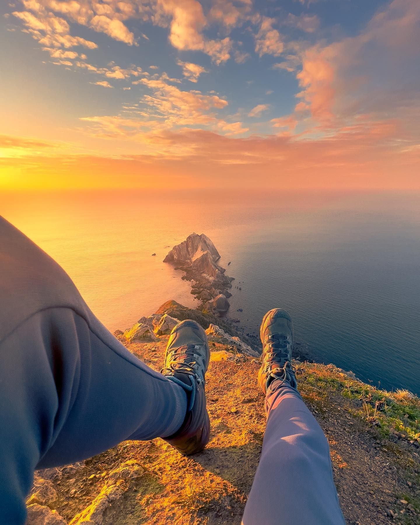 A person is sitting on a cliff overlooking the ocean at sunset.