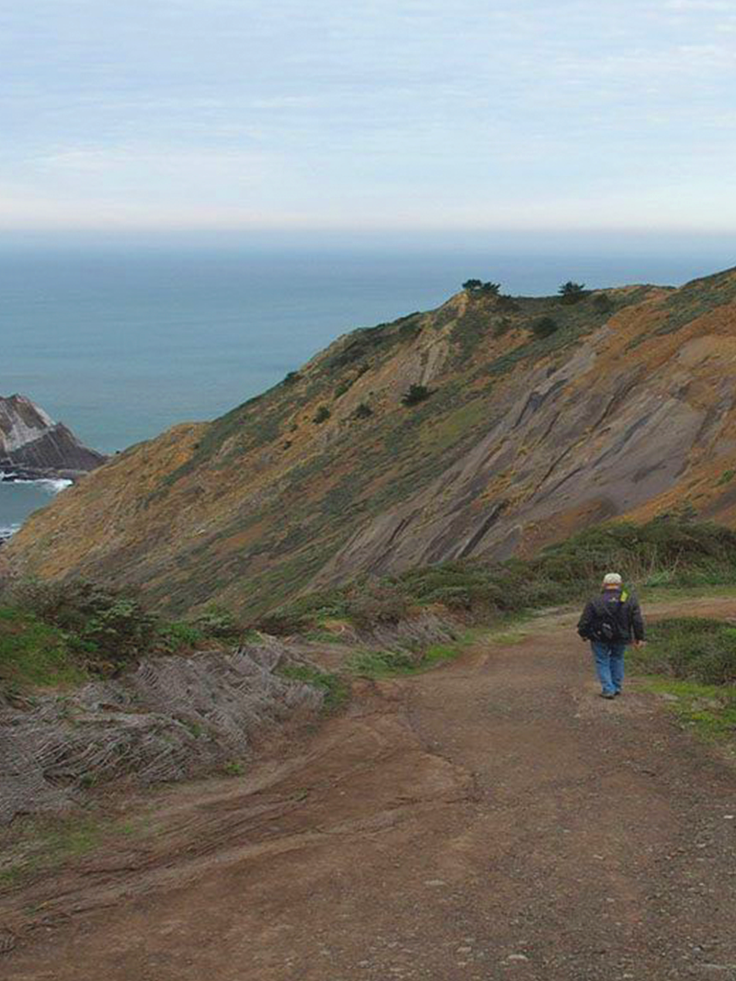 A man is walking down a dirt path on a hill overlooking the ocean.