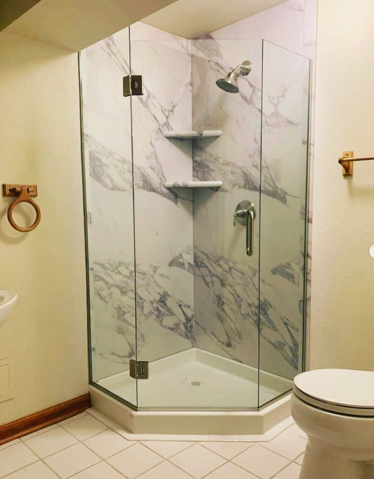 A marble-look corner shower with glass doors, shelves, and chrome fixtures. Toilet and towel rack visible.