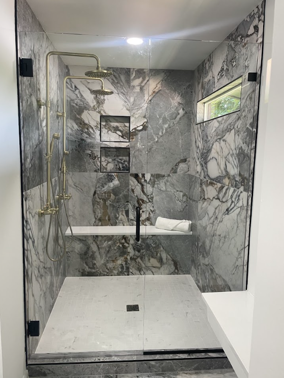 Modern marble-tiled walk-in shower with glass door, built-in shelf, and bench.